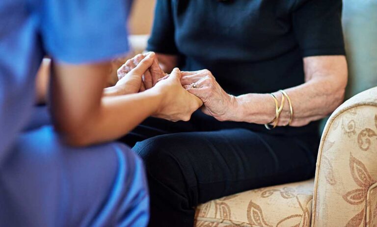 An elderly resident receives help with medication from a caregiver in a comfortable shared living space.